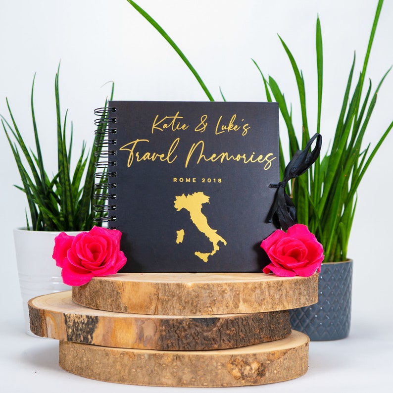 Peut inclure: Journal de voyage noir avec inscription dor&eacute;e "Katie & Luke's Travel Memories" et "Rome 2018". Le journal est pos&eacute; sur une pile de rondins de bois et entour&eacute; de plantes vertes.