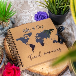 Peut inclure: Un journal de voyage marron avec une carte du monde noire et le texte "Adil & Orsi Travel memories" sur la couverture. Le journal a une reliure spirale noire et un ruban noir.