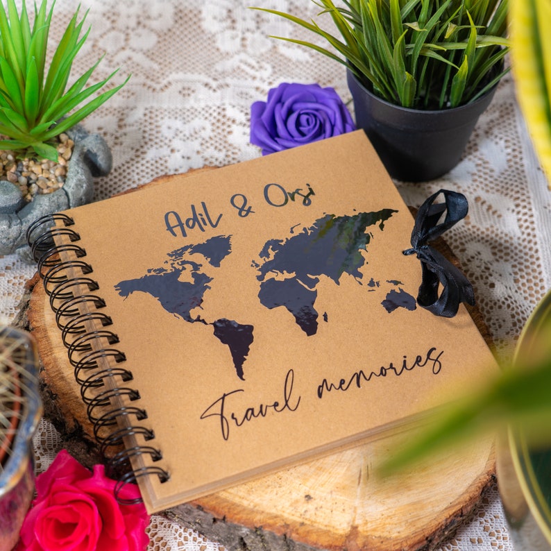 Peut inclure: Un journal de voyage marron avec une carte du monde noire et le texte "Adil & Orsi Travel memories" sur la couverture. Le journal a une reliure spirale noire et un ruban noir.