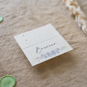 K&ouml;nnte beinhalten: Eine gefaltete, cremefarbene Karte mit dem Namen "Brianna" in Schreibschrift, verziert mit einem zarten blauen Blumendesign. Die Karte besteht aus strukturiertem Papier und ist wahrscheinlich eine Tischkarte oder Einladung.