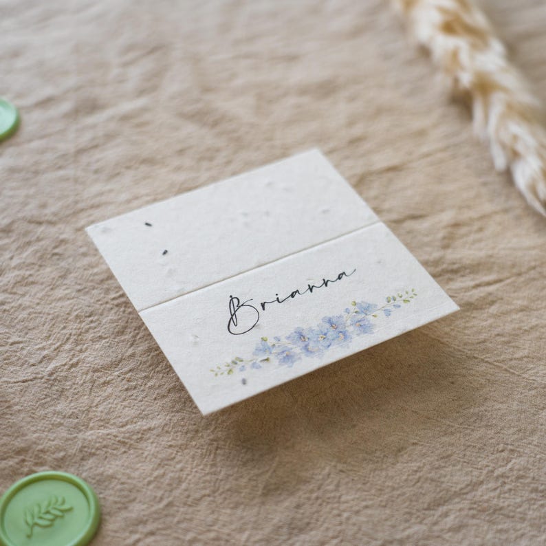 K&ouml;nnte beinhalten: Eine gefaltete, cremefarbene Karte mit dem Namen "Brianna" in Schreibschrift, verziert mit einem zarten blauen Blumendesign. Die Karte besteht aus strukturiertem Papier und ist wahrscheinlich eine Tischkarte oder Einladung.
