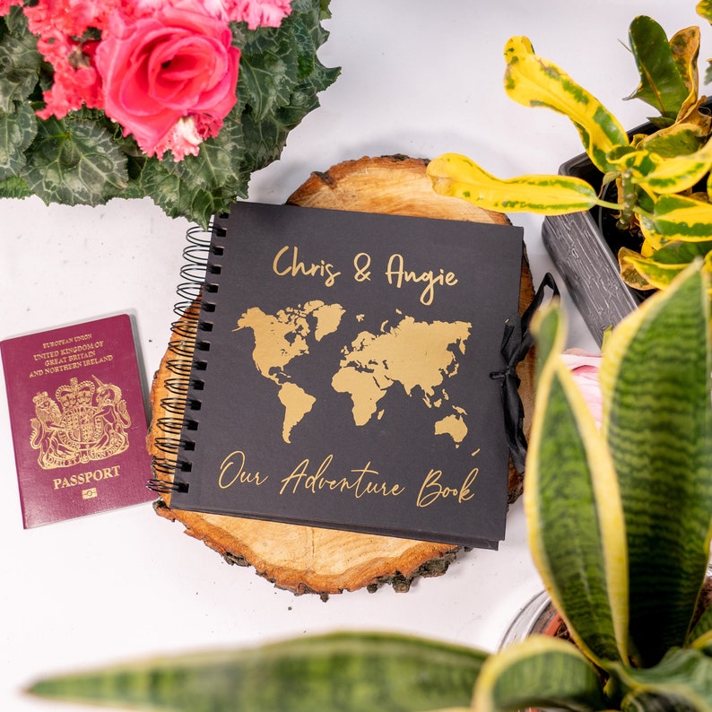 Peut inclure: Un journal reli&eacute; en cuir noir avec une inscription dor&eacute;e "Chris & Angie Our Adventure Book". Le journal est ouvert pour r&eacute;v&eacute;ler une carte du monde dor&eacute;e sur la premi&egrave;re page. Le journal est pos&eacute; sur une tranche de bois.