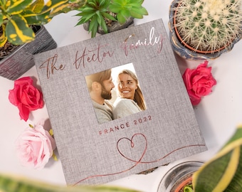 May include: A grey photo album with a linen texture and a rose gold heart design. The album has a photo window with a picture of a couple. The text "The Hector Family" and "France 2022" is printed in rose gold on the front of the album.