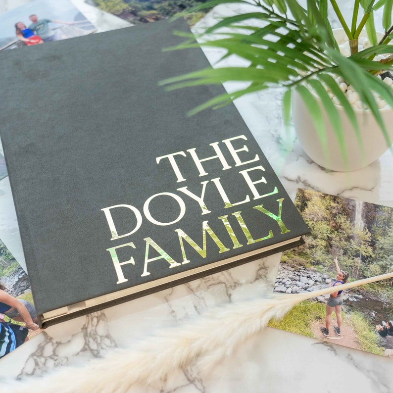May include: A dark gray hardcover photo album with the words THE DOYLE FAMILY printed in large, reflective gold lettering on the front cover. The album is positioned alongside several personal photographs and a small potted green plant.