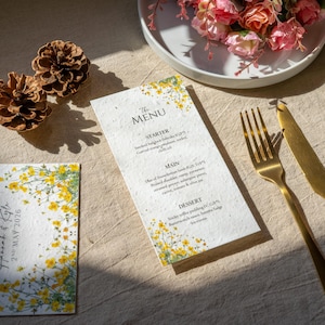 May include: A wedding menu and invitation card with floral accents. The menu features a starter, main course, and dessert. The invitation card displays the names "Hannah & Kyle" and the date "2nd May 2026". A gold fork and knife are on the table.
