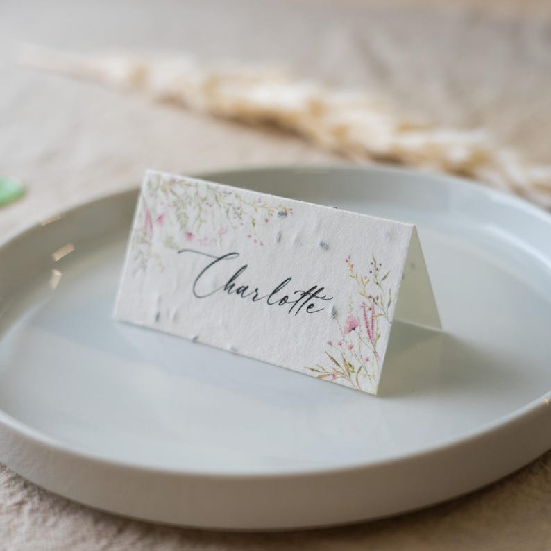 K&ouml;nnte beinhalten: Eine gefaltete Tischkarte mit dem Namen "Charlotte" in eleganter Schrift. Die Karte ist mit zarten floralen Illustrationen in Rosa, Gr&uuml;n und Lavendel verziert. Sie liegt auf einem wei&szlig;en Teller, was auf eine Hochzeits- oder Veranstaltungsdekoration hindeutet.