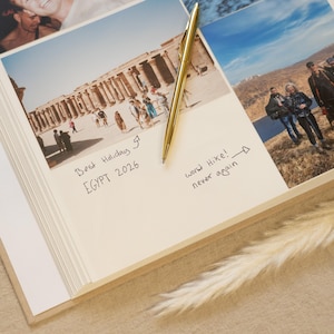 May include: A photo album open to a page with a photograph of an Egyptian temple and handwritten notes. A gold pen lies on the page. The text reads "Best Holiday Egypt 2026" and "Worst Hike! never again."
