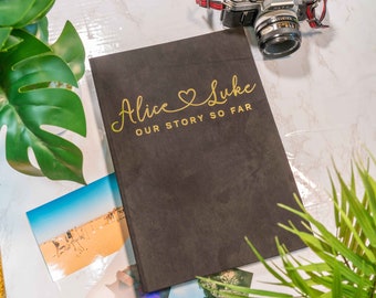 May include: A black photo album with the text "Alice & Luke Our Story So Far" in gold lettering. The album is on a white marble surface with a camera and green plants.