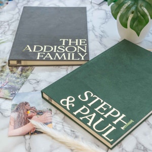 May include: Two personalised photo albums. One is dark grey with "THE ADDISON FAMILY" in gold lettering. The other is dark green with "STEPH & PAUL" in gold lettering. Photos are scattered around the albums.