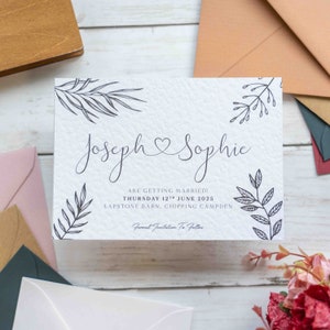 Op de afbeelding: Save the date-kaart met een witte achtergrond en zwarte inkt. De kaart heeft een eenvoudig bloemenontwerp en de tekst "Joseph & Sophie trouwen! Donderdag 12 juni 2025 Lapstone Barn, Chipping Campden Formele uitnodiging volgt"