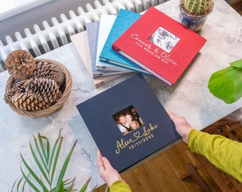 May include: A navy blue photo album with a gold inscription that reads "Alice & Luke 12/11/2022" and a small window showing a photo of a couple. The album is on a table with other photo albums in the background.