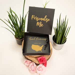 Peut inclure: Journal de voyage noir avec inscription dor&eacute;e "Personnaliser moi" et "Souvenirs de voyage Am&eacute;rique 2022". Le journal est ouvert pour r&eacute;v&eacute;ler un contour dor&eacute; des &Eacute;tats-Unis. Le journal est pos&eacute; sur une surface en bois avec des fleurs roses.