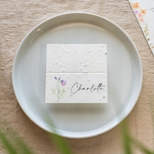 K&ouml;nnte beinhalten: Eine wei&szlig;e Tischkarte mit dem Namen "Charlotte" in eleganter Schrift, verziert mit einem Aquarell-Blumenmuster. Die Karte liegt auf einem wei&szlig;en Teller, passend f&uuml;r eine Hochzeit oder ein Event.