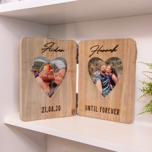 Puede incluir: Un marco de fotos de madera con dos aberturas en forma de corazón para fotos. El marco está dividido en dos secciones, cada una con un nombre y una fecha. El lado izquierdo dice "Aidan" y "21.08.20". El lado derecho dice "Hannah" y "UNTIL FOREVER".