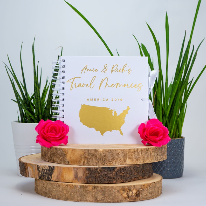 Peut inclure: Un carnet de voyage reli&eacute; en spirale blanc avec une inscription dor&eacute;e qui dit "Annie & Rich's Travel Memories" et "America 2019". Un contour dor&eacute; des &Eacute;tats-Unis est sur la couverture. Le carnet est pos&eacute; sur trois rondins de bois.