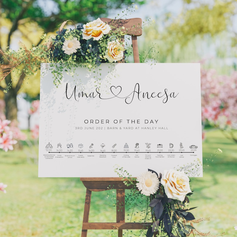 May include: A white wedding sign with the names "Umar" and "Aneesa" in cursive script, a heart symbol, and the text "ORDER OF THE DAY" with the date "3RD JUNE 202" and the location "BARN & YARD AT HANLEY HALL". The sign also includes a timeline of wedding events with icons and times.
