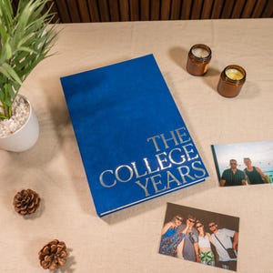 May include: A blue photo album with silver text reading "THE COLLEGE YEARS". Two photographs and two small candles are placed near the album. A small plant and pine cones are also on the table.