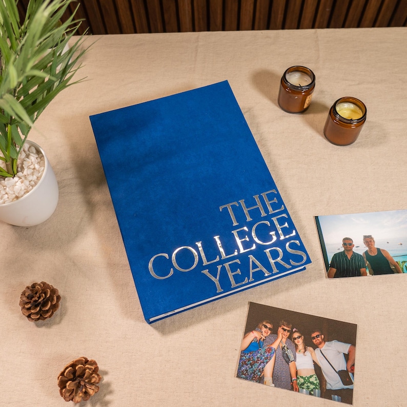 May include: A blue photo album with silver text reading "THE COLLEGE YEARS". Two photographs and two small candles are placed near the album. A small plant and pine cones are also on the table.