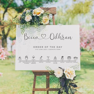 May include: A white wedding sign with the names Becca and Odhran in cursive script, a heart symbol, and the text "Order of the Day" with the date and location. The sign lists the wedding day schedule with icons and times for each event.
