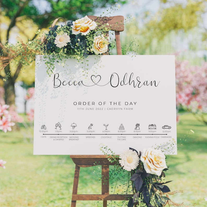 May include: A white wedding sign with the names Becca and Odhran in cursive script, a heart symbol, and the text "Order of the Day" with the date and location. The sign lists the wedding day schedule with icons and times for each event.