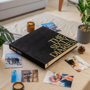 May include: A square black photo album with gold lettering on the cover that reads THE SIXTY DIARIES and the date 18.09.1961 on the spine. The album is surrounded by several loose photographs, a small amber glass candle jar, and a pinecone.