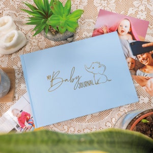 Puede incluir: Un diario para beb&eacute; de color azul claro con una ilustraci&oacute;n de elefante dorada y el texto "My Baby Journal".