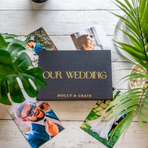 Peut inclure: Un album photo noir avec l'inscription dorée "OUR WEDDING" et les noms "HOLLY & CRAIG". Des photos de mariage et de la verdure entourent l'album sur une surface en bois blanc.