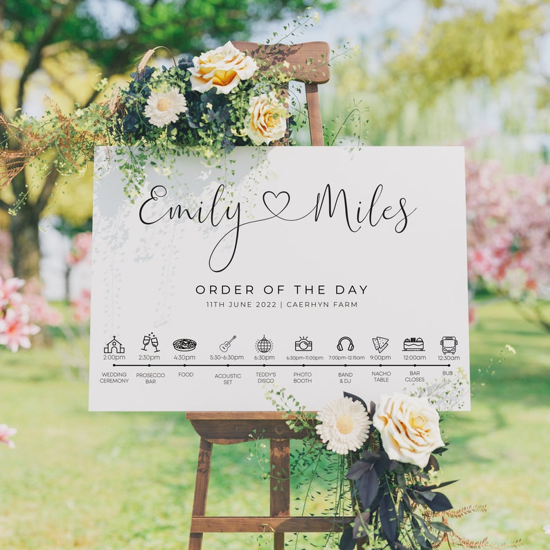 May include: A white wedding sign with black text and icons. The sign reads "Emily & Miles Order of the Day 11th June 2022 | Caerhyn Farm". The sign lists the wedding day schedule with times and activities.