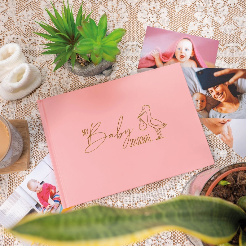 Puede incluir: Un diario de beb&eacute; rosa con una ilustraci&oacute;n de cig&uuml;e&ntilde;a dorada y el texto "My Baby Journal".