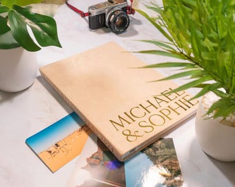May include: A light-colored wooden photo album with the names "MICHAEL & SOPHIE" in gold lettering. A vintage camera with a red strap sits above the album. Several photos are scattered around the album, and two potted plants are in the background.