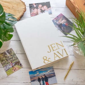 Może przedstawiać: Biały album fotograficzny z pozłacanym napisem "Jen & Avi". Album leży na białym drewnianym blacie, wokół niego rozrzucone są zdjęcia.
