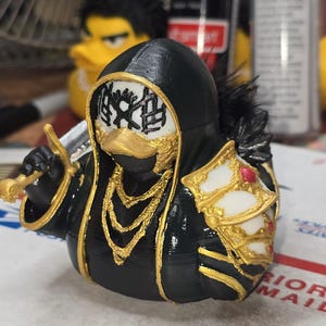 May include: A black rubber duck figurine dressed as a warrior in a hooded cloak with gold trim, a gold chain necklace, and a white face mask featuring a black sun-like symbol. The duck holds a silver sword and wears a white and gold shield on one side.
