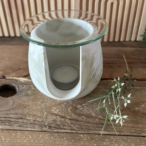 Könnte beinhalten: Ein weiß-grüner, marmorierter Diffusor für ätherische Öle mit einer durchsichtigen Glasschale. Eine brennende Teelichtkerze befindet sich im Inneren. Der Diffusor steht auf einer Holzoberfläche mit dekorativem Grün.