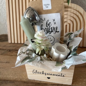Könnte beinhalten: Eine Holzkiste mit den Worten "Glücksmomente" und einem Herzsymbol. Die Kiste enthält einen Teebeutel, ein Tee-Ei aus Metall, eine grüne Kerze, eine weiße Blume und einen blütenförmigen weißen Kerzenhalter. Auf einer weißen Karte steht "be happy".