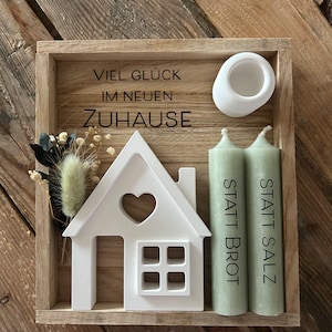 Könnte beinhalten: Eine Holzkiste mit einem weißen Haus-Ausschnitt, zwei grünen Kerzen und einem weißen Kerzenhalter. Die Kiste trägt den Text "Viel Glück im neuen Zuhause" und "Statt Salz Statt Brot".