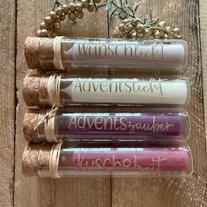 Könnte beinhalten: Vier Glasröhrchen mit Korkstopfen, die jeweils eine farbige Kerze enthalten. Die Röhrchen sind mit goldener Schrift versehen, die Wunschlicht, Adventslicht, Adventszauber und Kuschelzeit lautet. Ein dekorativer Zweig mit goldenen Beeren liegt darüber.
