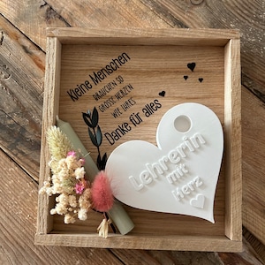 May include: A wooden box with a white heart-shaped plaque that says "Lehrerin mit Herz" (Teacher with heart). The box also has a small bouquet of dried flowers and a green and white rolled up paper.