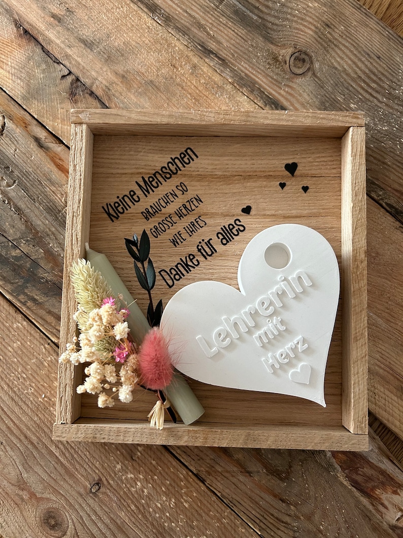 May include: A wooden box with a white heart-shaped plaque that says "Lehrerin mit Herz" (Teacher with heart). The box also has a small bouquet of dried flowers and a green and white rolled up paper.