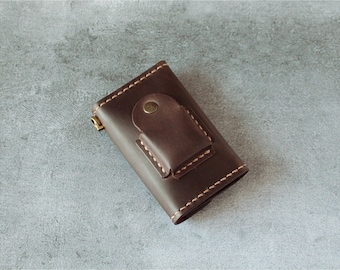 May include: A dark brown leather key holder with a flap closure secured by a brass button. The edges are stitched with light-colored thread. A small brass zipper is visible on the side.