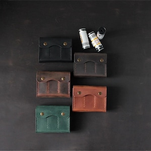 May include: Five rectangular leather cases in black, dark brown, light brown, and green, each featuring a flap with two brass snap buttons and vertical stitching. Three cylindrical film canisters are positioned near the top right case.