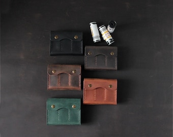 May include: Five rectangular leather cases with flap closures secured by two brass-colored metal snaps. The cases are arranged in black, dark brown, medium brown, light brown, and green colors, accompanied by three cylindrical film canisters.