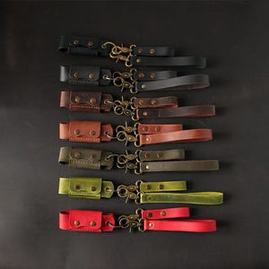May include: A collection of leather wrist straps in various colors, including black, brown, and red. Each strap features a small pouch with snap closures and a brass-colored clasp. The straps are arranged on a dark surface.