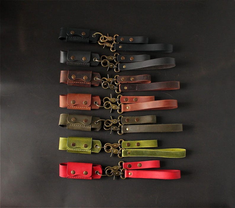 May include: A collection of leather wrist straps in various colors, including black, brown, and red. Each strap features a small pouch with snap closures and a brass-colored clasp. The straps are arranged on a dark surface.