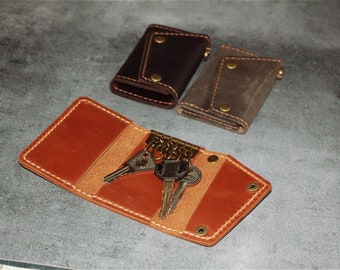 May include: Three leather keychains with different colored leathers. The keychain on the left is brown, the keychain in the middle is gray, and the keychain on the right is orange. The orange keychain has a set of keys attached to it.
