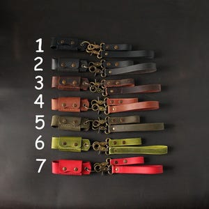 May include: A collection of seven leather wrist straps in various colors, including black, brown, green, and red. Each strap features a small pouch, metal hardware, and a matching wrist loop. The straps are arranged against a dark background.