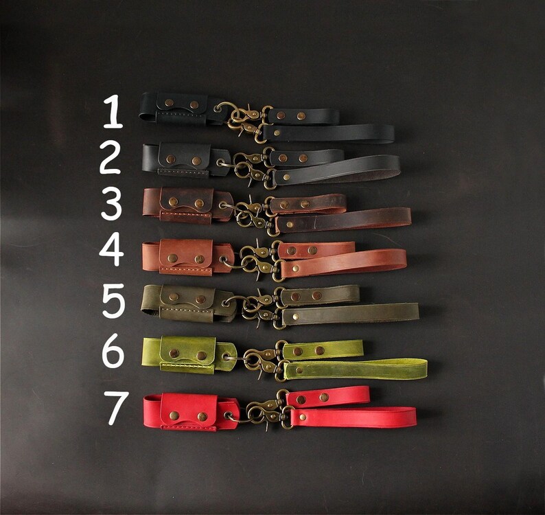 May include: A collection of seven leather wrist straps in various colors, including black, brown, green, and red. Each strap features a small pouch, metal hardware, and a matching wrist loop. The straps are arranged against a dark background.