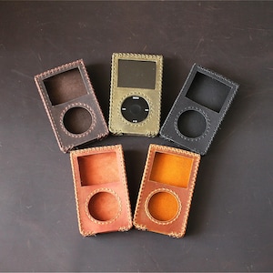 May include: Five rectangular leather cases for portable media players arranged in a fan shape. The cases come in dark brown, olive green, black, reddish-brown, and tan. Each case features a square screen cutout, a circular click wheel cutout, and visible stitched edges.