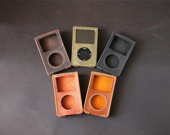 May include: Five rectangular leather cases for portable media players arranged in a fan shape. The cases come in dark brown, olive green, black, reddish-brown, and tan. Each case features a square screen cutout, a circular click wheel cutout, and visible stitched edges.