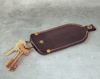 May include: A dark brown leather key holder with a stitched border and brass rivets. Two keys are attached to a metal ring, partially inserted into the holder. The key holder is on a gray surface.