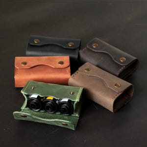 May include: A collection of five rectangular leather cases with snap closures, including black, tan, dark brown, and green options. One green case is open, revealing three cylindrical film canisters stored inside the main compartment.